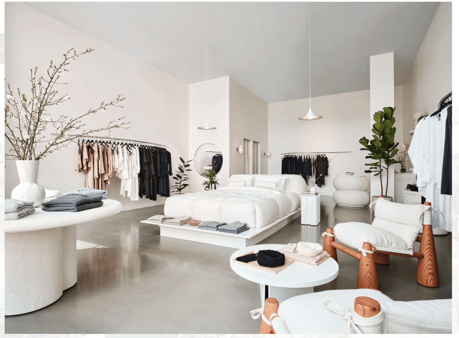 Bright minimalist boutique featuring a central white bed, neutral clothing racks, and modern white furniture.